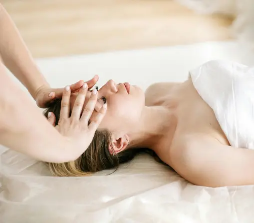 Therapist giving a relaxing head massage to a woman lying on a spa bed, wrapped in a white sheet, in a peaceful spa setting.